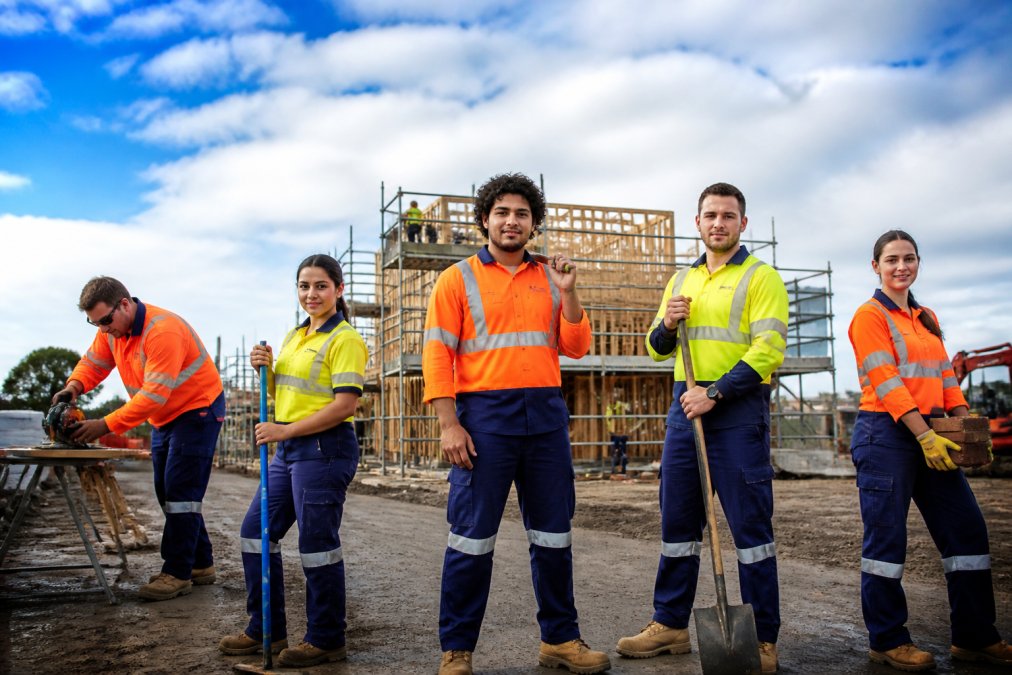 A diverse construction team wearing high-visibility workwear in different fabric compositions on an active job site, showing practical workwear in use across multiple trades with varying weather conditions, from sunny to partly cloudy