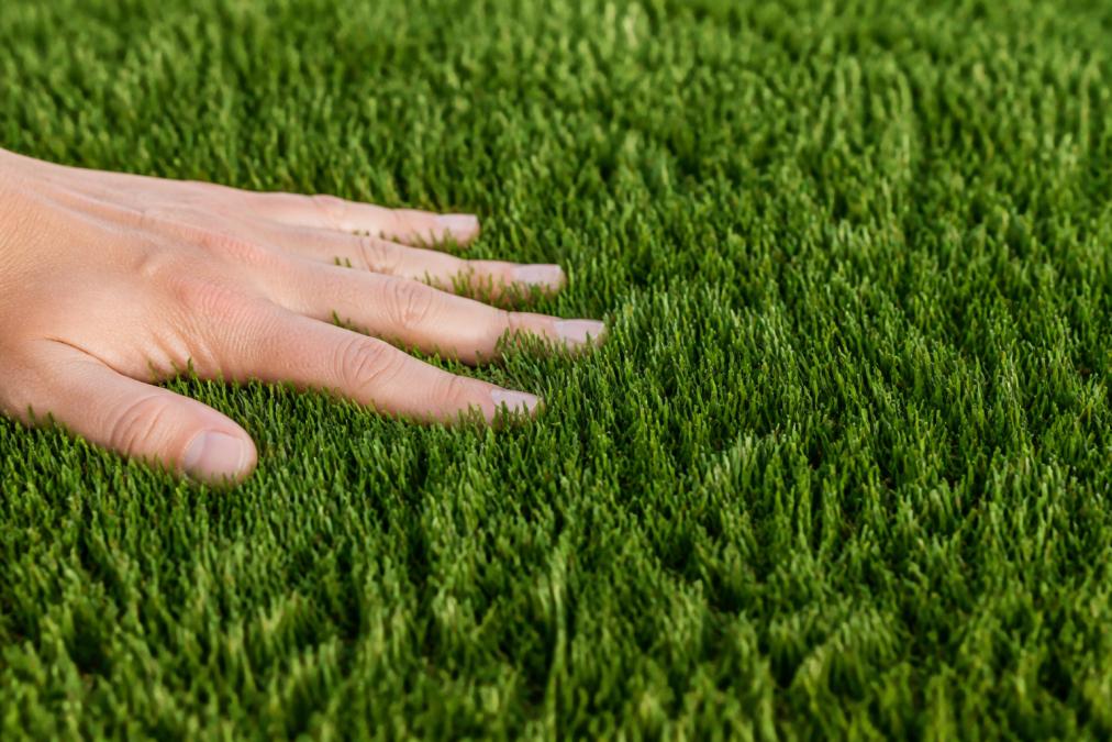 Close-up of polyethylene artificial grass fibers showing soft texture and natural green color variation, with a hand touching the surface to demonstrate softness