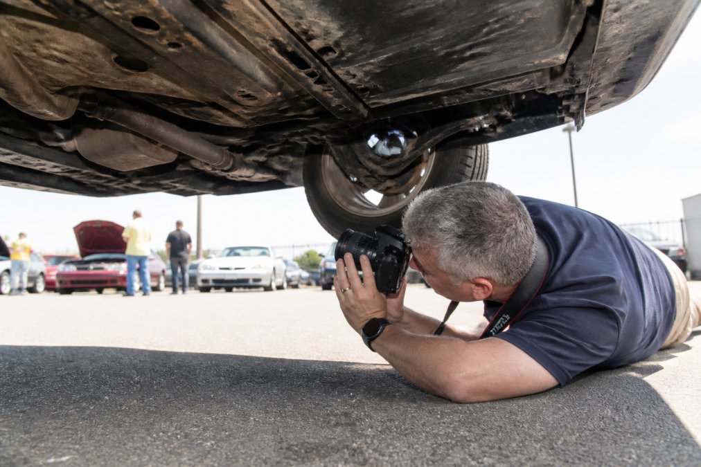 Professional mobile inspector conducting detailed vehicle examination at an outdoor auction lot, photographing undercarriage with professional camera, natural daylight