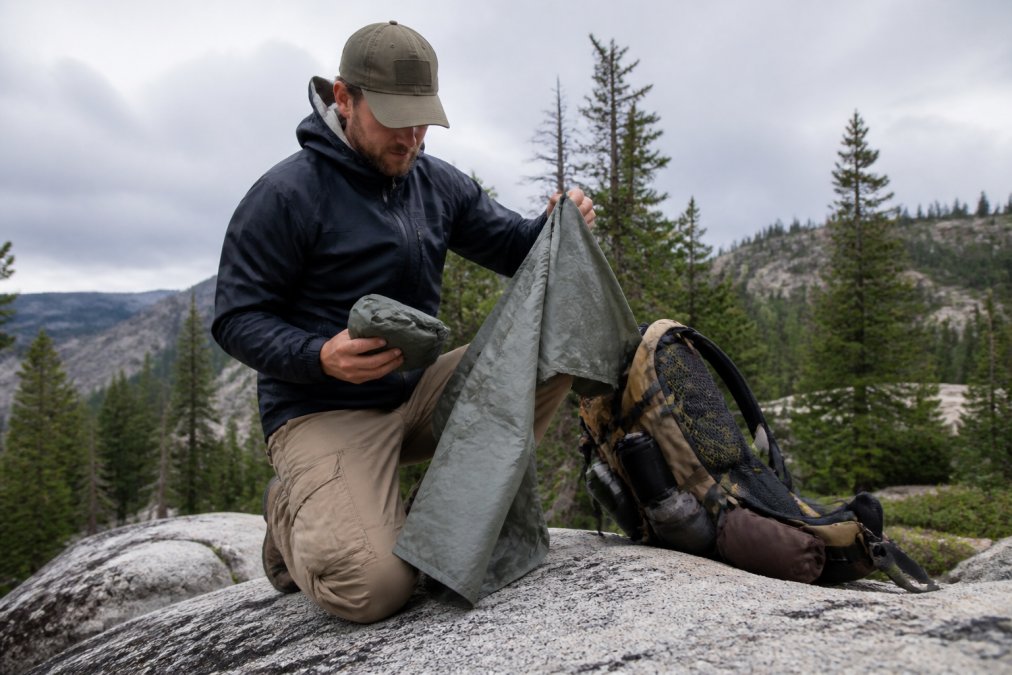 Hiker kneeling on a granite outcrop unfolding a packable poncho from its stuff sack, with a loaded backpack and pine forest behind, soft overcast light