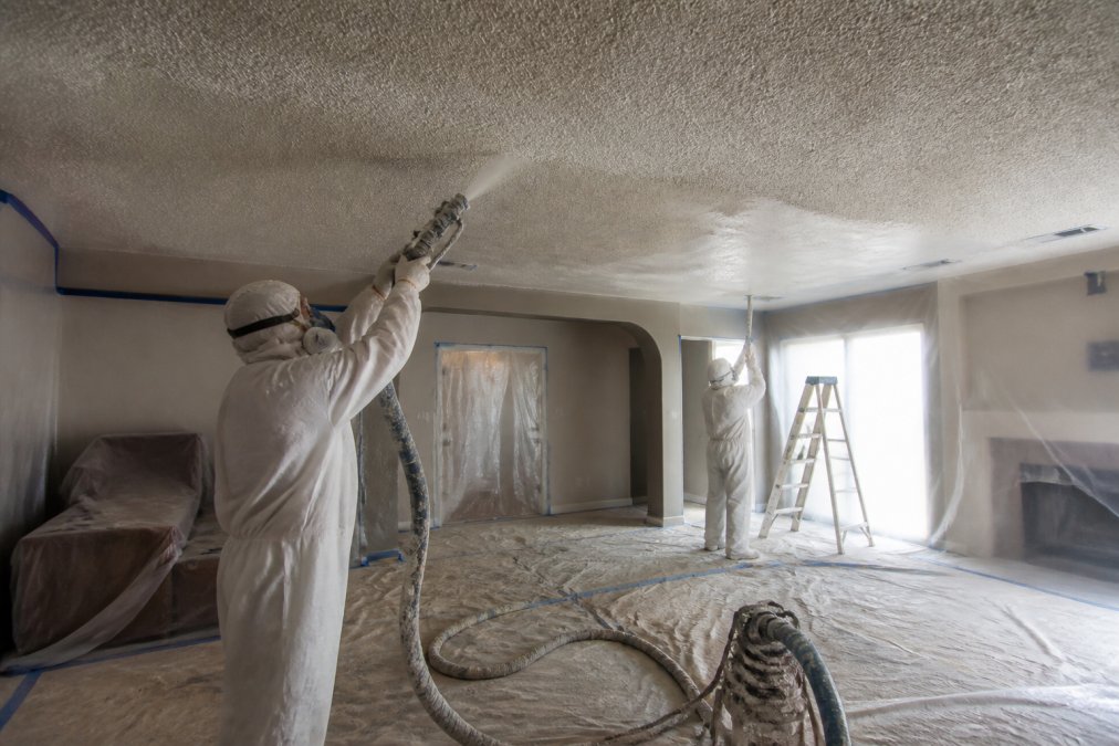 Professional crew in safety gear and drop cloths carefully removing popcorn texture from ceiling with spray equipment, showing proper protection and workspace setup