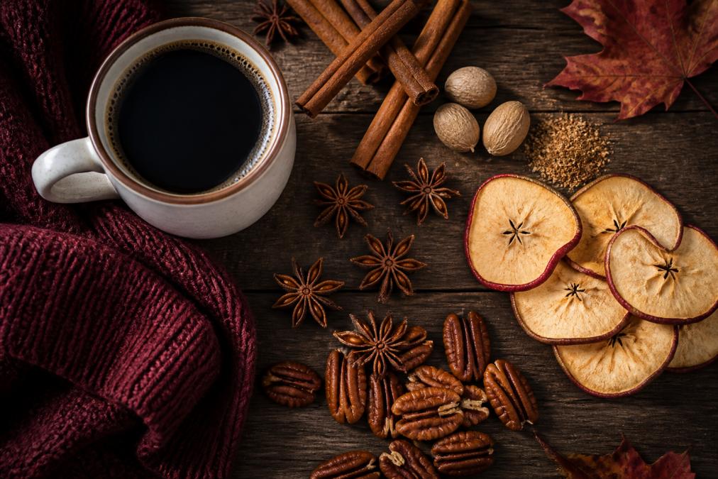 Overhead shot of seasonal coffee ingredients arranged on a rustic wooden surface: cinnamon sticks, star anise, nutmeg, dried apple slices, and pecan halves next to a warm mug of coffee