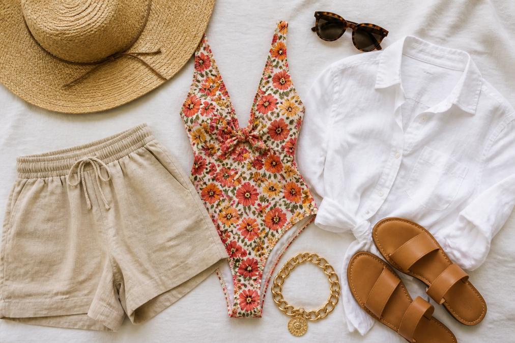 A flat-lay of neatly arranged vacation clothing on a white linen surface, a bright floral swimsuit, a white linen button-down shirt, tan linen shorts, tan leather sandals, oversized tortoiseshell sunglasses, a wide-brimmed straw hat, and a chunky gold statement necklace arranged in a cohesive coastal palette