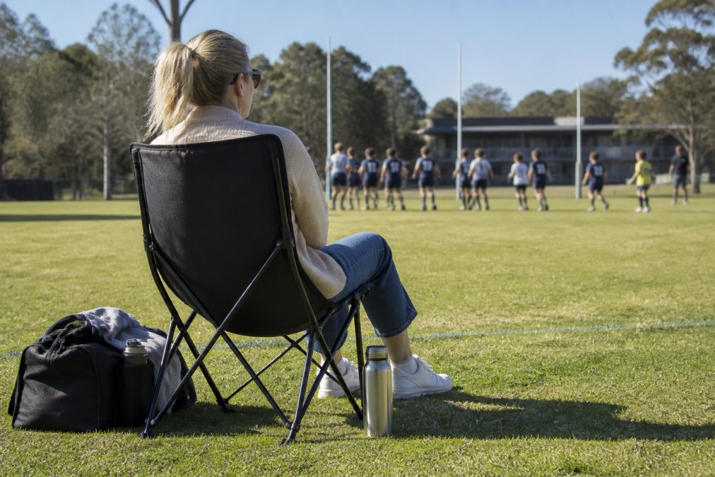 A parent sitting in a compact black folding chair on the sideline of a junior footy oval on a sunny weekend, with a sports bag and water bottle beside the chair and kids training in the background