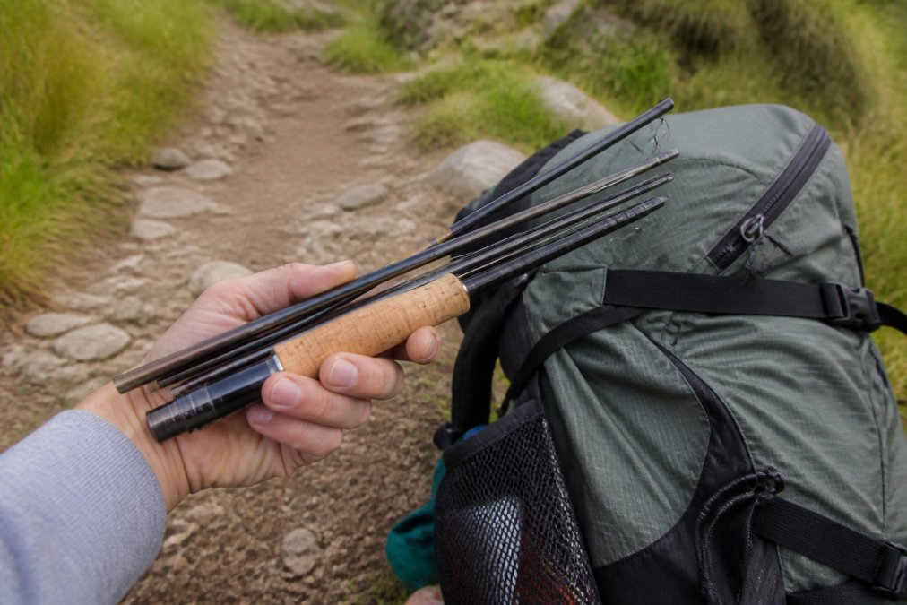 Close-up of a hand holding a collapsed multi-piece fly rod next to a hiking backpack on a dirt trail, showing the compact size for travel