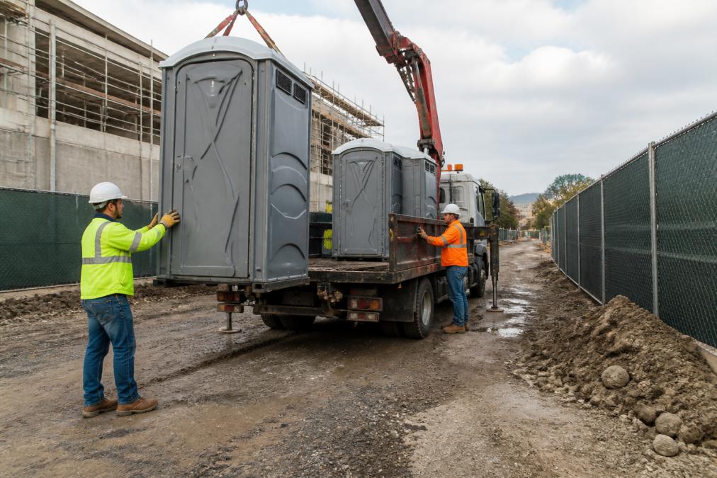 A delivery truck positioning a portable restroom on an active construction site, with workers in the background and temporary fencing lining the perimeter