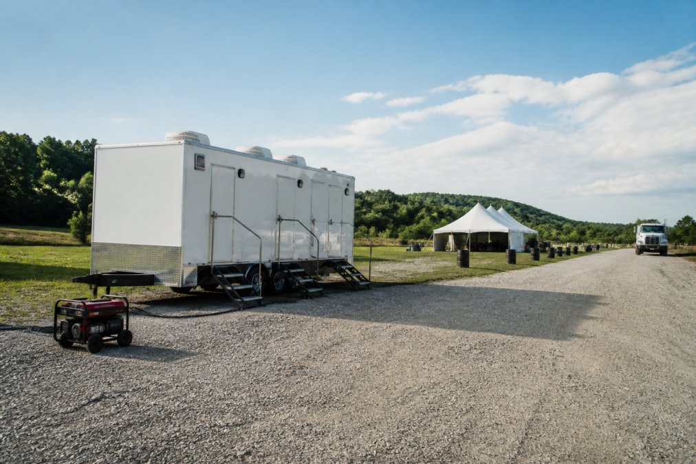 A white luxury restroom trailer positioned on flat gravel ground at an outdoor event site, with a visible power cord running along the ground to a portable generator nearby, and a clear access path wide enough for a delivery truck, photographed in afternoon sunlight