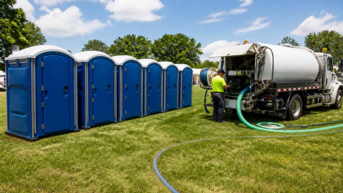 A row of clean portable restrooms set up on a grassy field at an outdoor event venue, blue sky overhead, a white service truck parked nearby with hoses and supplies visible