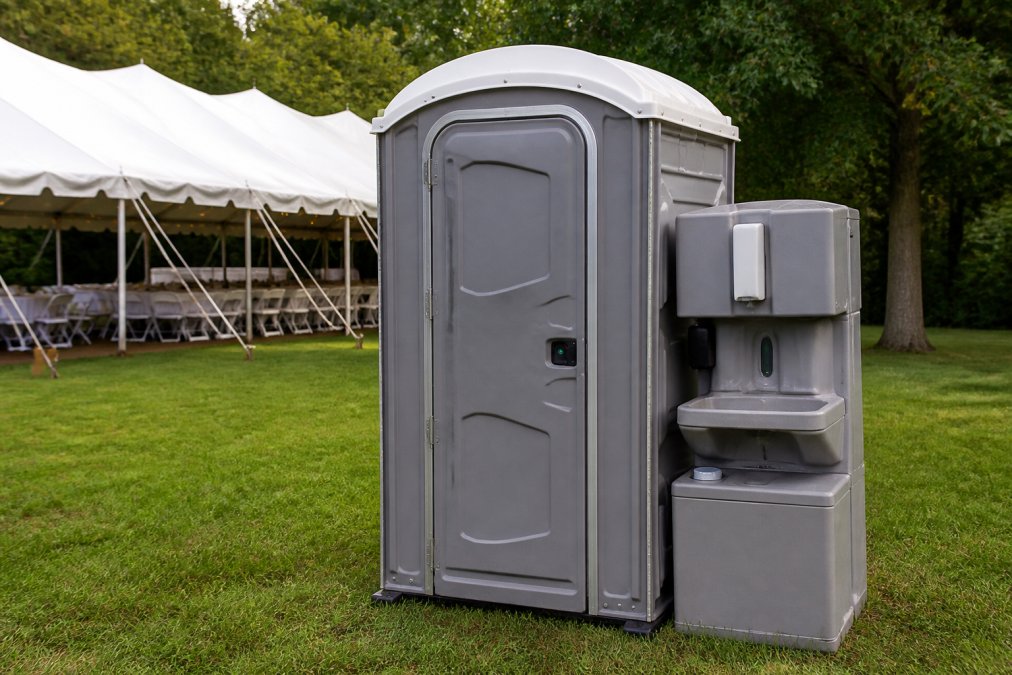 Professional portable restroom unit with handwashing station attached, positioned on grassy event venue with clear signage visible, natural daylight illuminating the clean exterior