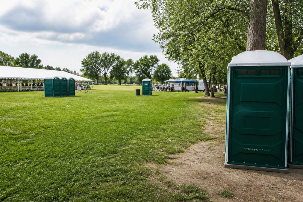 Outdoor event venue with multiple portable restroom units strategically spaced around grassy area, clear signage visible, adequate distance maintained from guest seating and food service areas, natural daylight showing clean setup