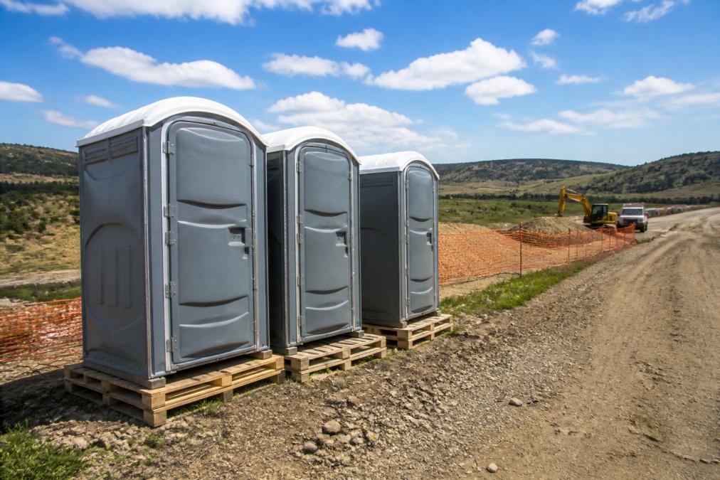 Rural construction site with portable toilets positioned on wooden pallets for ground stability, showing proper spacing between units and access road, with rolling hills and open terrain in background