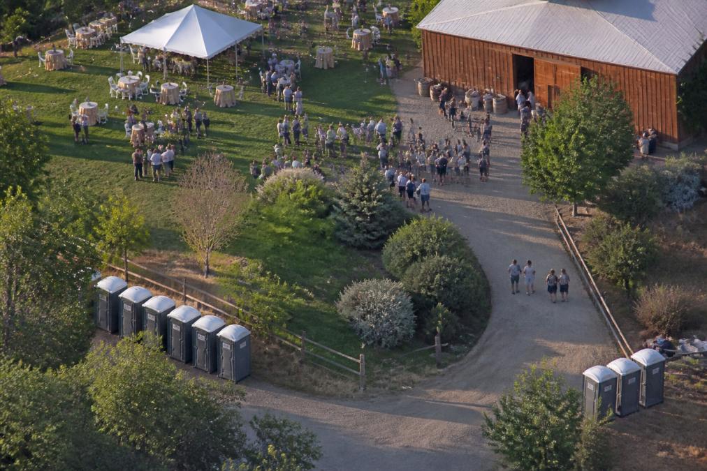 Overhead view of outdoor event venue with multiple portable restroom units strategically positioned around perimeter, clusters of attendees visible in background during daytime event