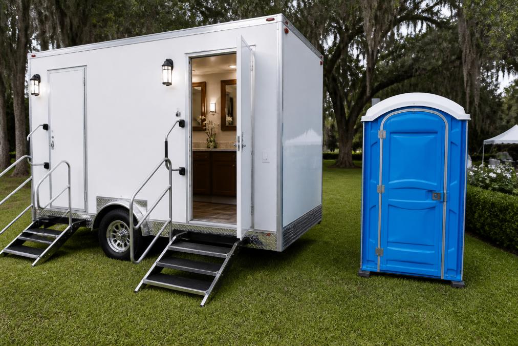 A clean, well-lit luxury restroom trailer parked on manicured grass at an outdoor wedding reception, door open to reveal wood-trimmed mirrors, vanity countertops, and decorative laminate flooring, with a standard blue portable toilet unit visible in the background
