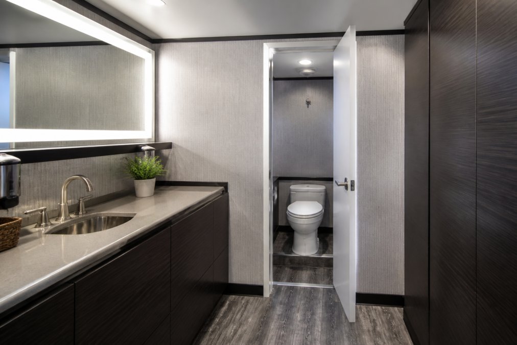 Interior of a premium restroom trailer showing climate-controlled space with decorative laminate flooring, LED-lit vanity mirror, freshwater sink with polished fixtures, and a private flushable toilet stall with a clean white door