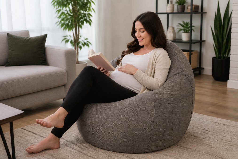 Femme enceinte souriante assise confortablement dans un pouf ergonomique en tissu gris dans un salon moderne et lumineux, une main posée sur son ventre et l'autre tenant un livre