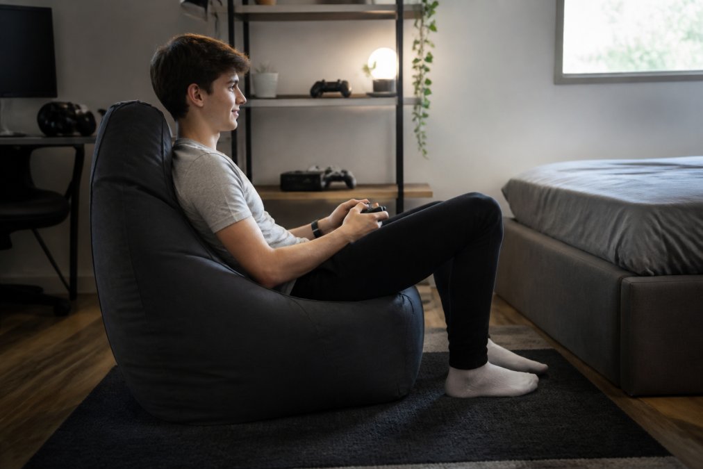 Gamer in ergonomic beanbag pouf with proper lumbar support, leaning back comfortably with console controller in hands, showing aligned spinal posture during extended gaming session in modern bedroom