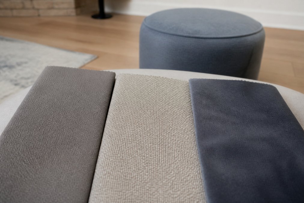 Gros plan de plusieurs échantillons de tissus (microfibre lisse, polyester à grain fin, velours ras) disposés côte à côte sur une surface neutre, montrant clairement la texture et la densité de chaque matériau, avec un petit pouf en arrière-plan