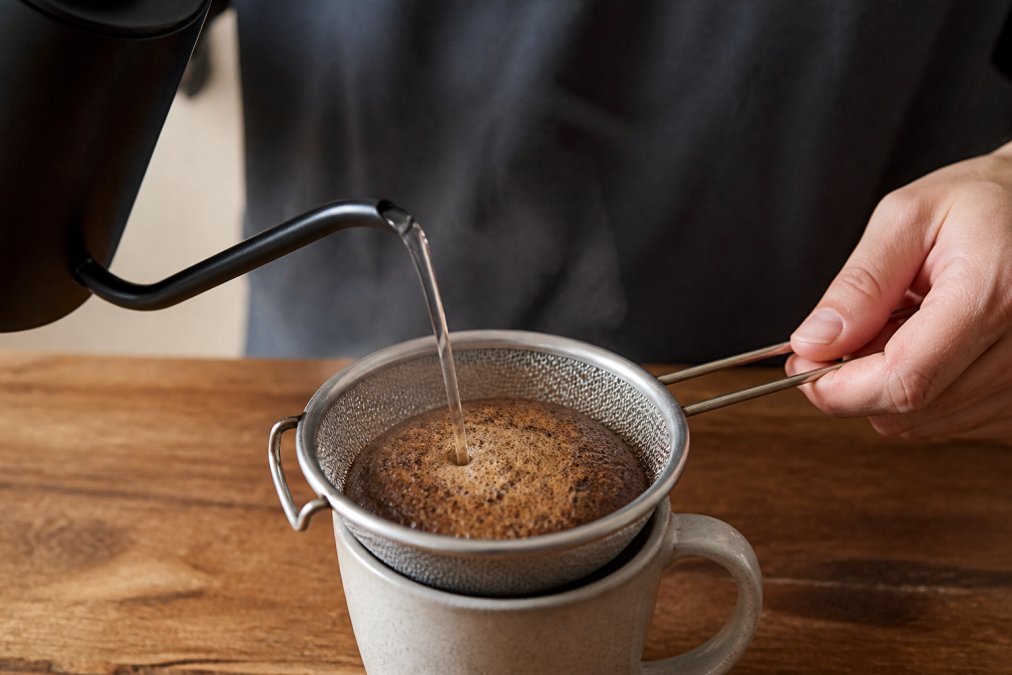 Close-up of hands pouring hot water over coffee grounds in a makeshift dripper made from a metal mesh strainer, with steam rising visibly, showing the bloom phase in action over a ceramic mug