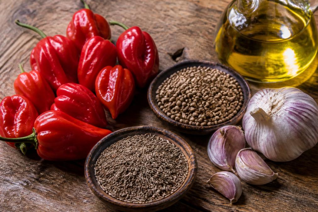 Gros plan de piments habanero rouges frais, graines de carvi, graines de coriandre, gousses d'ail et filet d'huile d'olive disposés sur une surface en bois rustique avec lumière naturelle