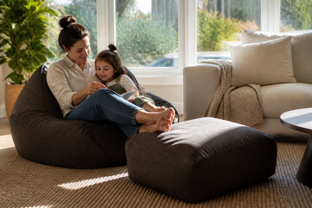 Parent and child relaxing together on a comfortable pouf in a bright living room, with the pouf serving as both seating and a footrest, natural afternoon sunlight streaming through windows
