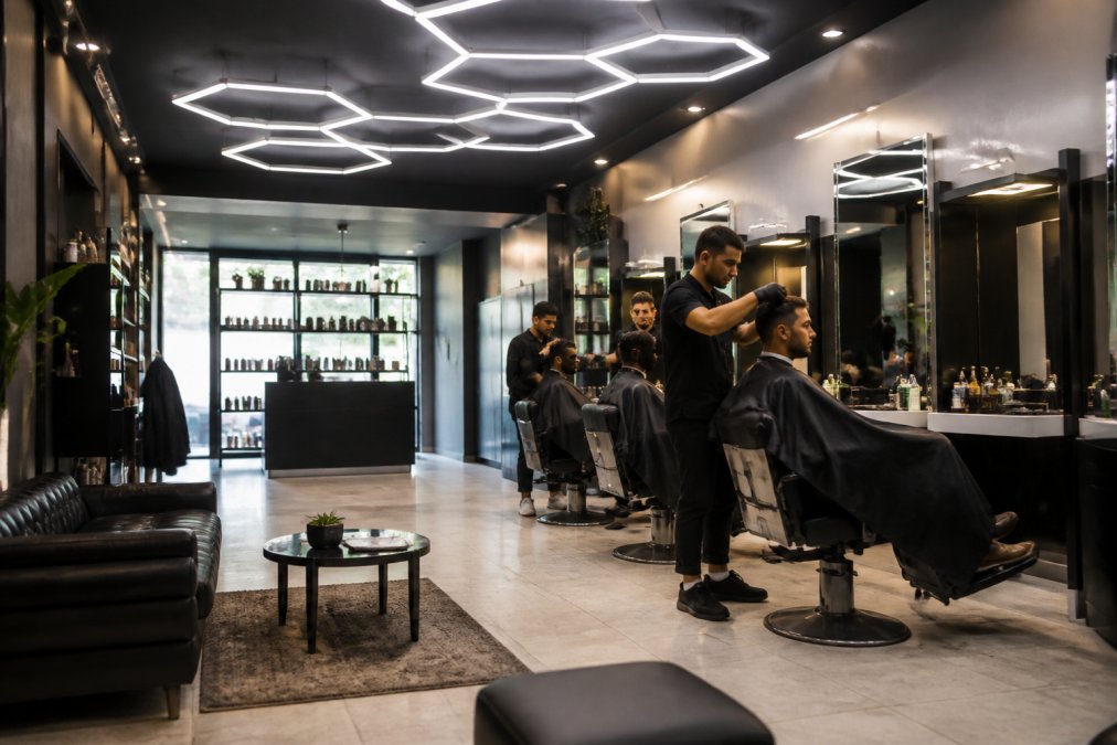 Modern, clean barbershop interior with comfortable seating, professional hexagonal lighting, and welcoming atmosphere with clients receiving services