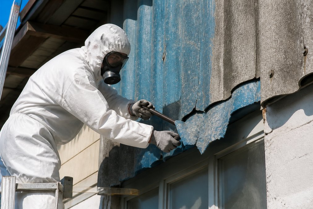 Certifikovaný pracovník v bílém ochranném obleku s respirátorem a brýlemi bezpečně demontující modrý azbestový materiál z fasády budovy v slunečním dni