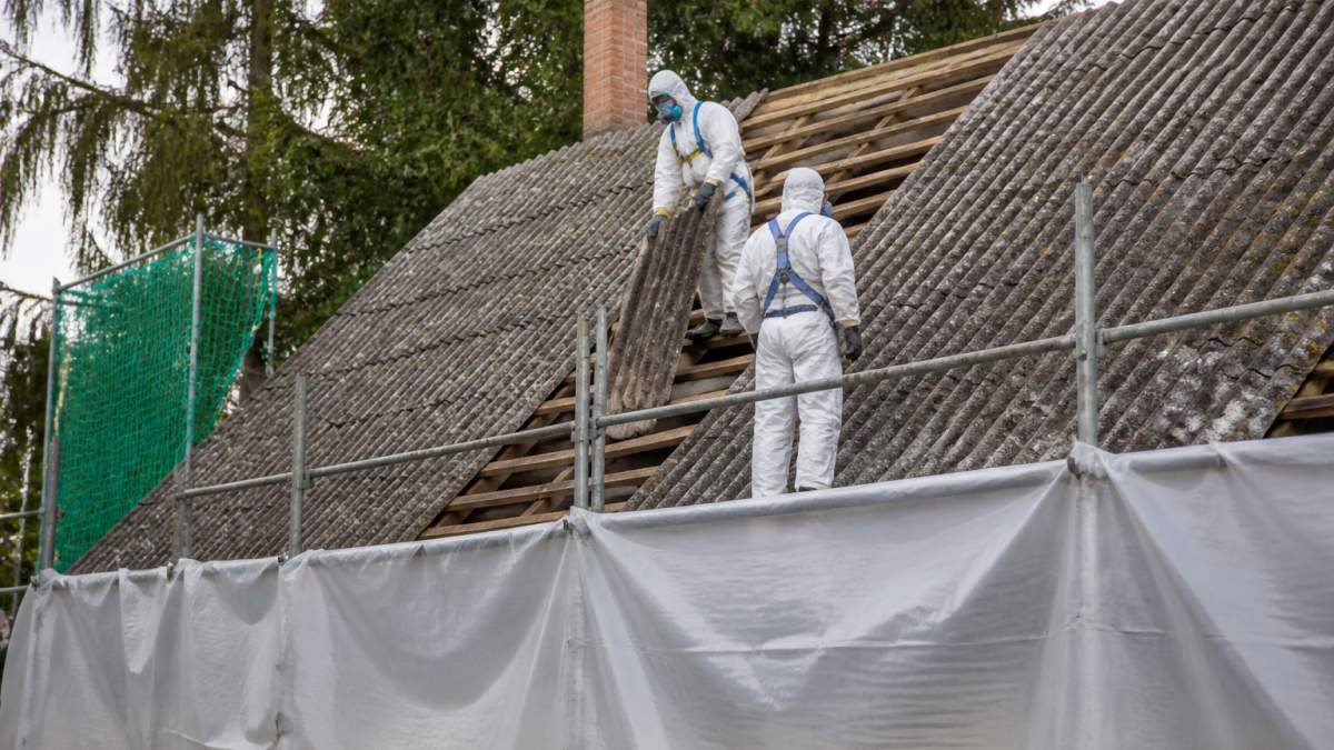 Pracovníci v ochranných oblecích a respirátorech demontují eternitové šablony ze střechy rodinného domu, kolem je zajištěno zábradlí a plachta proti pádu materiálu