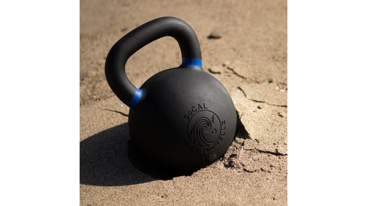 Close-up of a person's hands gripping a worn, rusting weight plate beside a new powder-coated competition kettlebell, contrasting surface textures under bright gym lighting