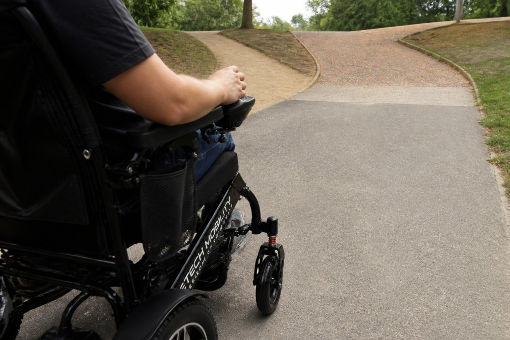 Close-up of a user's hands on the powerchair controls in an outdoor setting with varied terrain visible ahead including slight inclines and flat surfaces