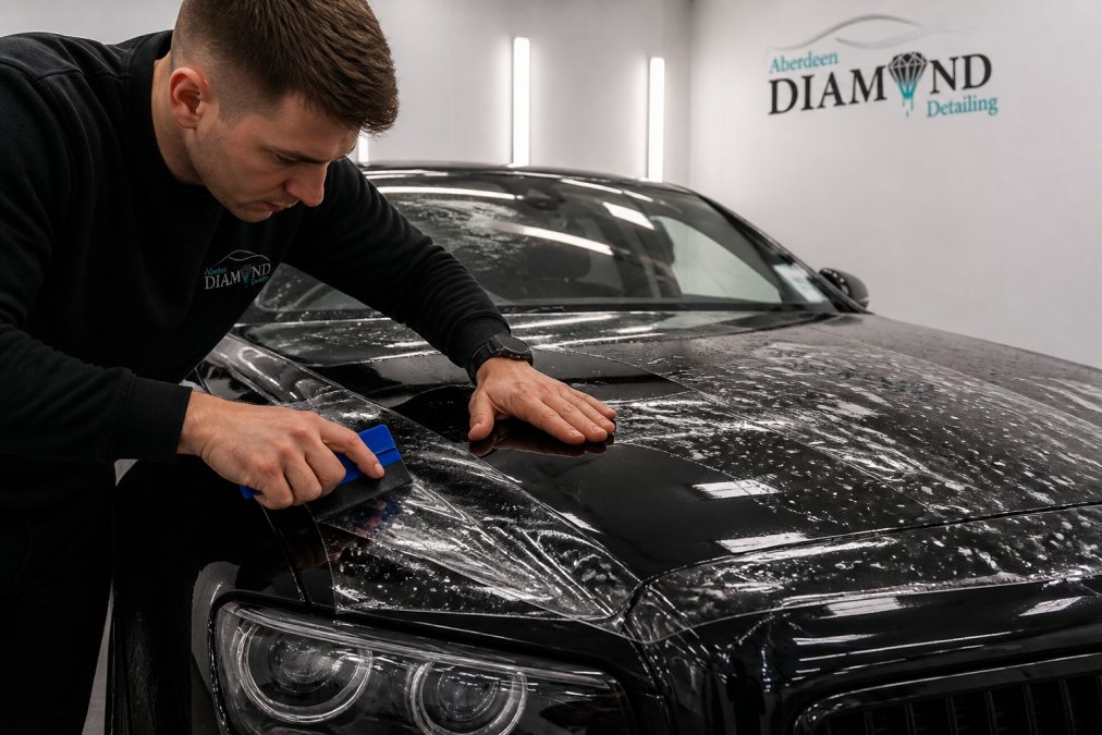 A detailer's hands smoothing transparent paint protection film across the bonnet of a dark prestige car with a squeegee, clean workshop lighting overhead