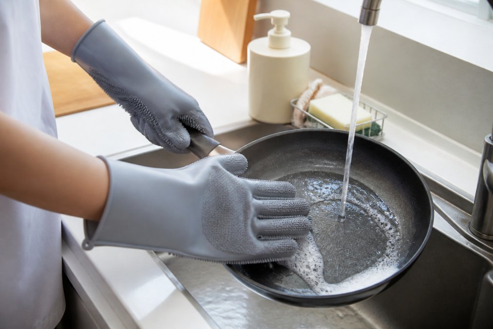 Close-up of hands wearing textured silicone scrubbing gloves washing a pan at a clean kitchen sink, with a soap dispenser and sponge rack visible in the background, bright natural light