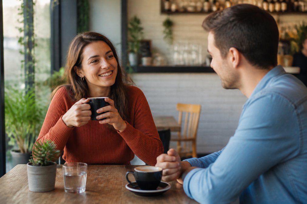 Two people engaged in a relaxed, natural conversation in a casual café setting, showing comfortable body language, genuine smiles, and easy interaction