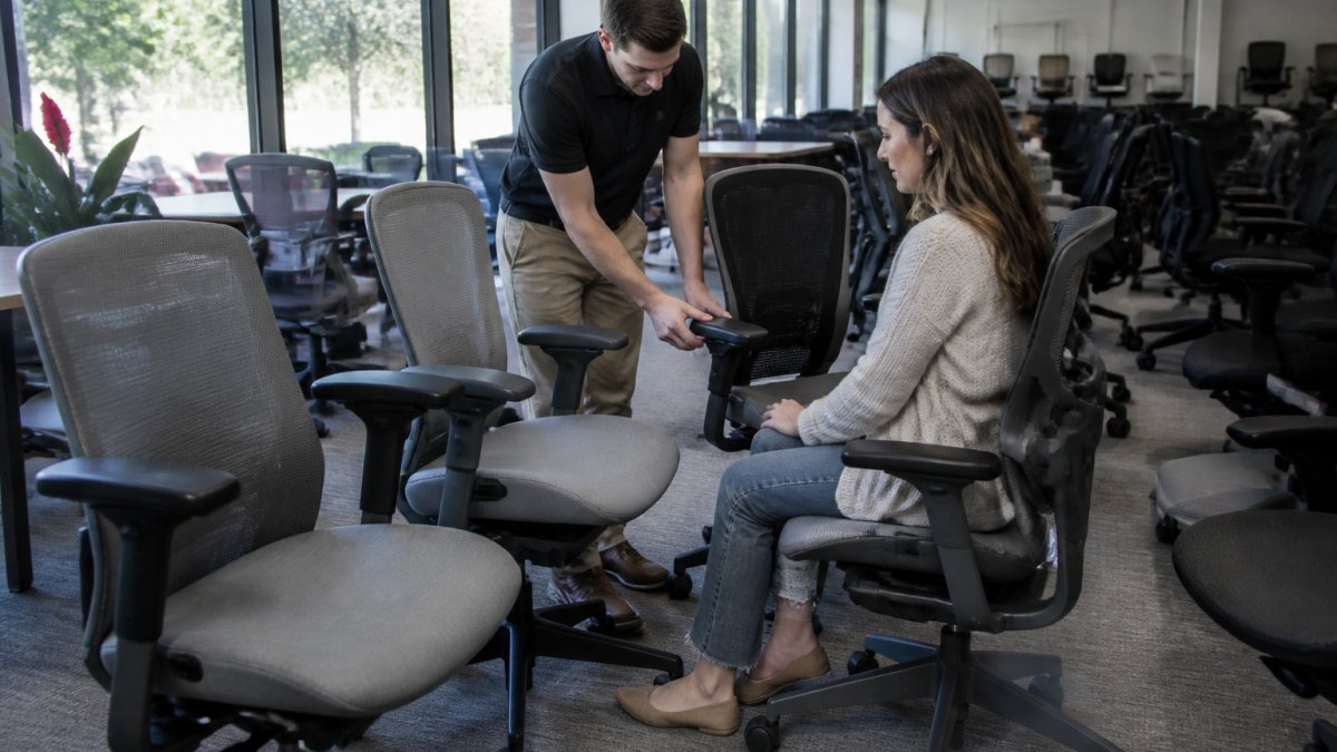 A well-lit furniture showroom with rows of pre-owned Steelcase office chairs in gray and black, a salesperson demonstrating the adjustable armrest to a customer, large windows letting in natural light