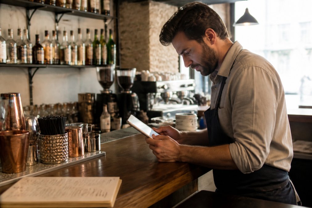 Dueño de restaurante pequeño revisando precios de software en una tablet mientras está detrás de la barra, con menú en papel visible al fondo