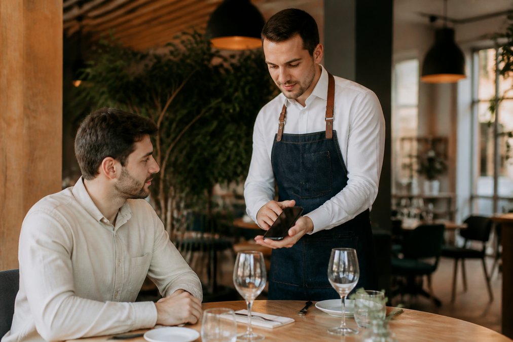 Camarero en un restaurante moderno mostrando un menú digital en un smartphone a un cliente sentado en la mesa, con iluminación cálida y ambiente profesional