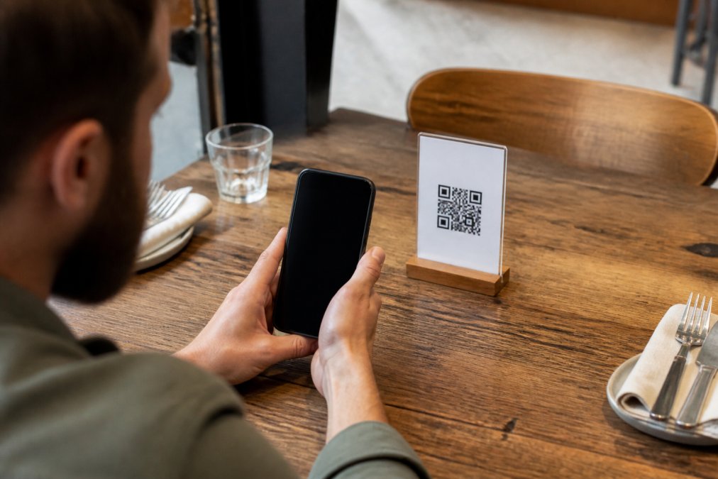 Cliente de un restaurante escaneando un código QR con su móvil para acceder al menú digital, con la pantalla del smartphone visible mostrando el menú cargado