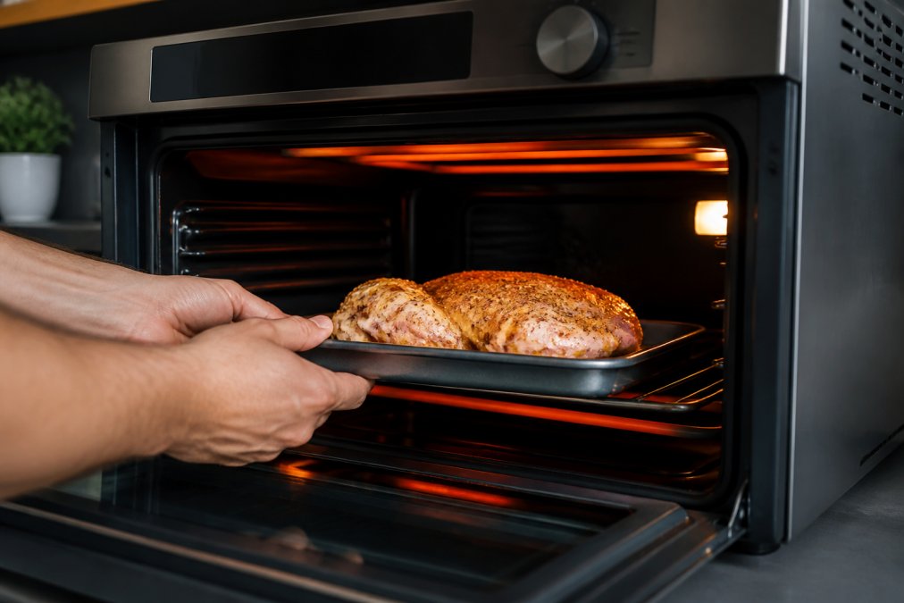 Close-up of hands placing seasoned chicken into a modern countertop oven with glowing heating elements and digital temperature display visible