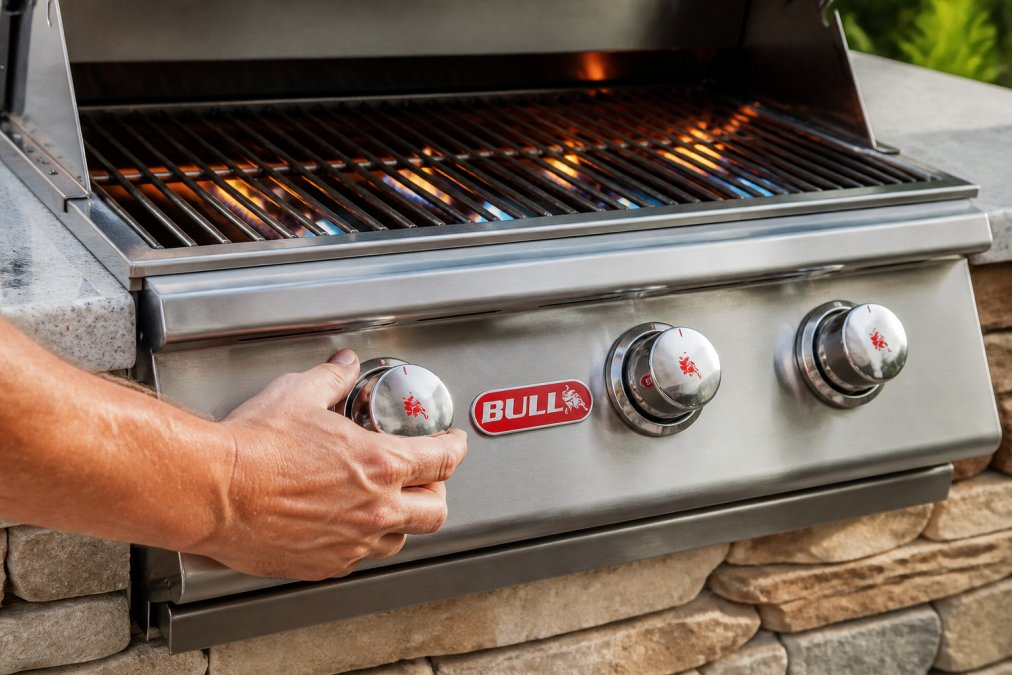 Close-up of a homeowner's hand adjusting stainless steel burner knobs on a gas grill with flames visible beneath the cooking grates, adjusting for optimal temperature control