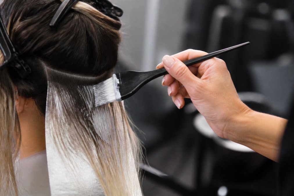 Professional colorist's hand holding a precision tint brush with feathered synthetic bristles, demonstrating proper grip and bristle detail during active hair coloring application on sectioned hair