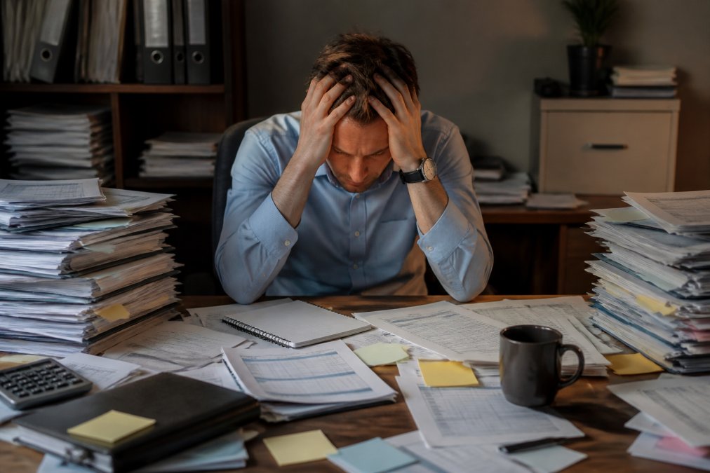 A sales manager at a cluttered desk looking overwhelmed by spreadsheets, sticky notes, and scattered paper reports in a dimly lit office