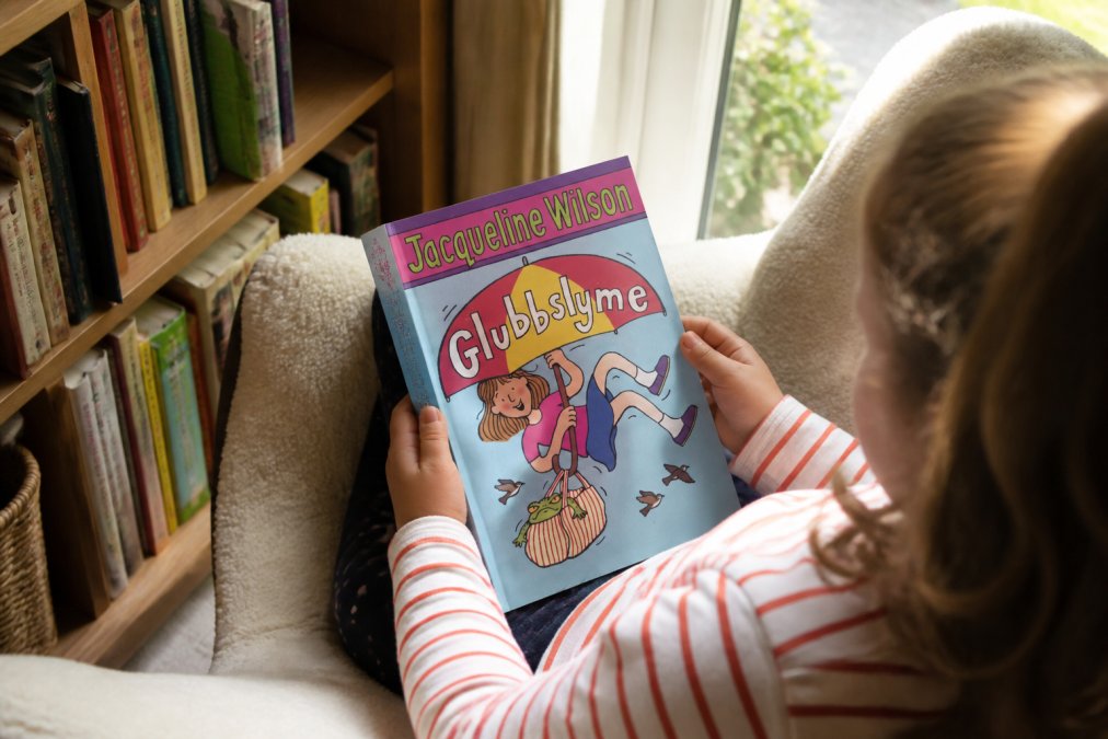 Child's hands holding open a colourful preloved children's book with bright illustrations, sitting on a cosy reading chair with natural daylight streaming through a window