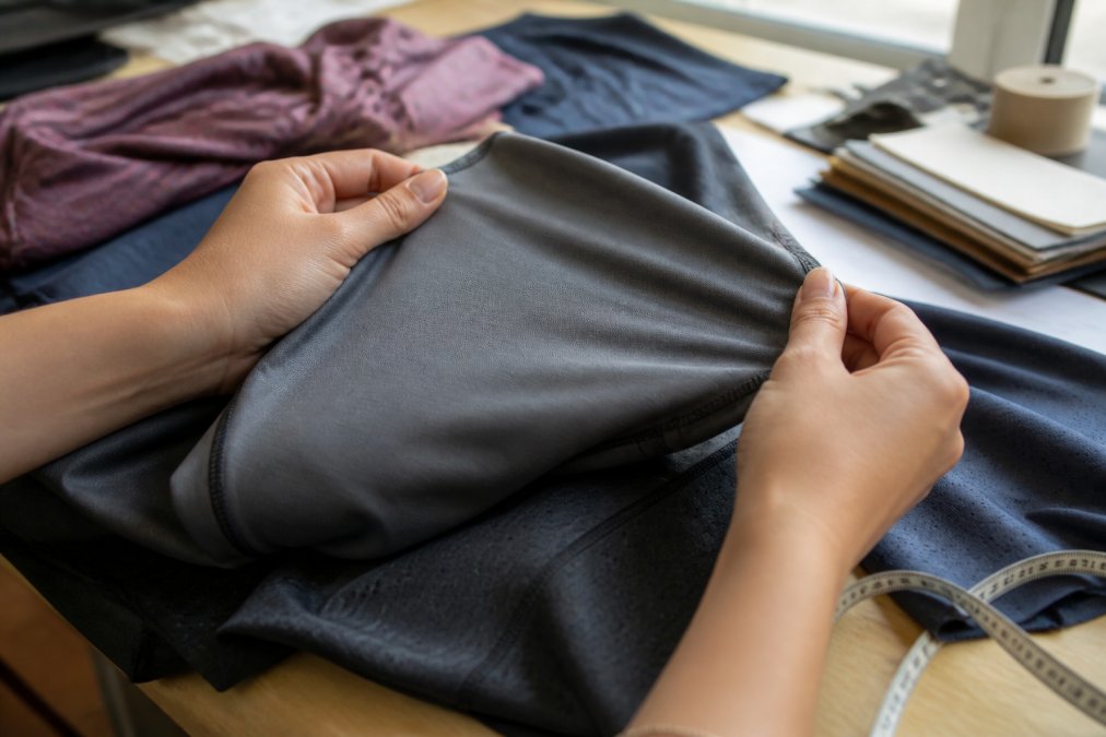 Close-up of hands testing the stretch and elasticity of premium athletic fabric, fingers pinching the weave to examine thickness and recovery, natural daylight illuminating the textile detail