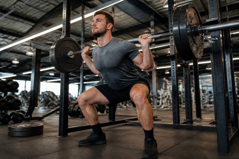 Man wearing premium athletic shorts performing a weighted squat in a modern gym with bright overhead lighting, demonstrating fabric stretch and fit during intense lower-body training