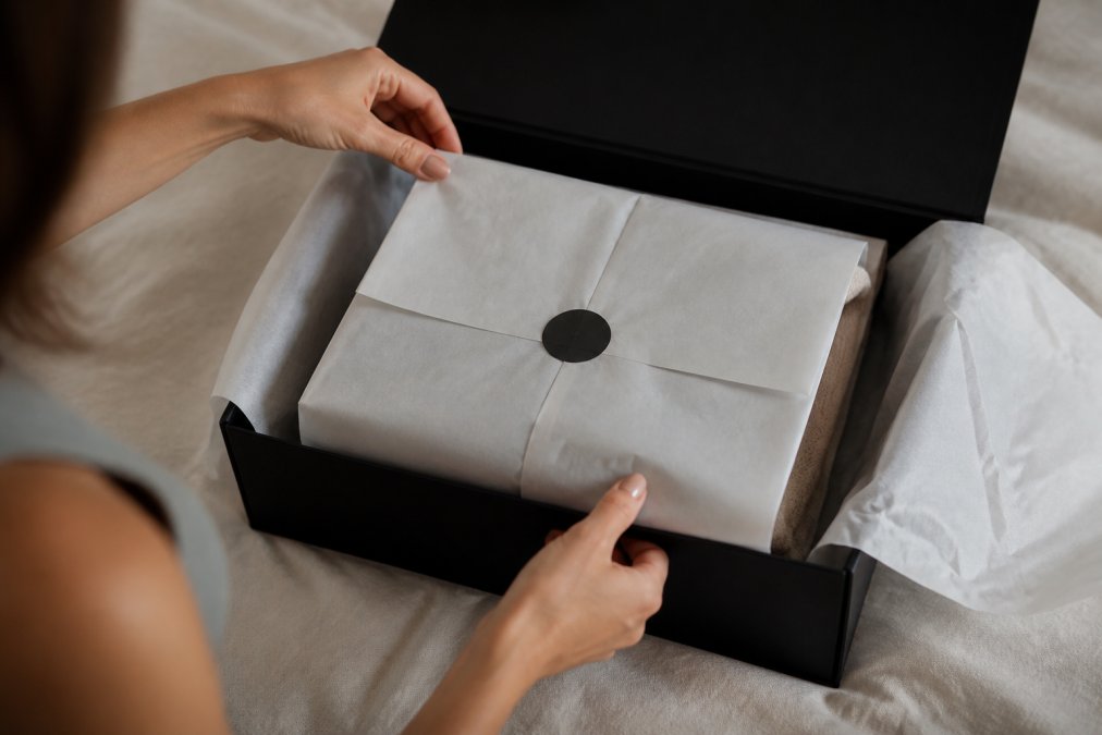 Hands carefully opening a luxury apparel box with layered tissue paper and branded elements visible, showing premium unboxing with soft natural lighting and elegant presentation