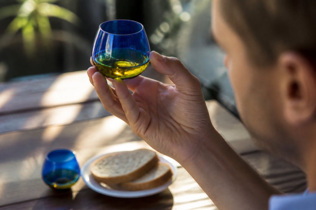 Close-up of a person holding a small blue tasting glass of golden green olive oil up to the light, with a slice of bread and a second glass on a wooden table