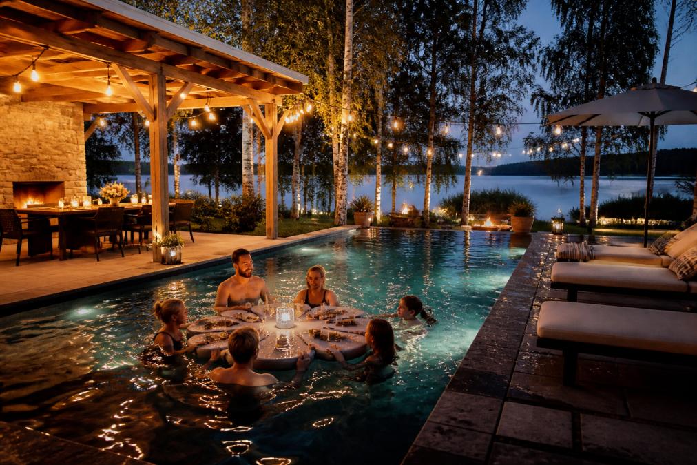 An evening backyard pool party scene: adults and children in a pool with a floating game table holding drinks, sun loungers and a pergola with a dining area alongside the pool, warm ambient lighting strung overhead, Finnish lakeside garden setting with birch trees in the background