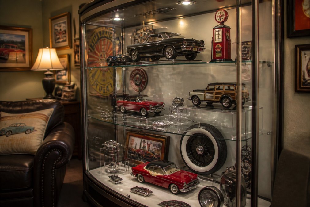 A well-lit glass display cabinet in a home den, featuring a classic car model, vintage signs, and neatly arranged memorabilia on tiered acrylic risers