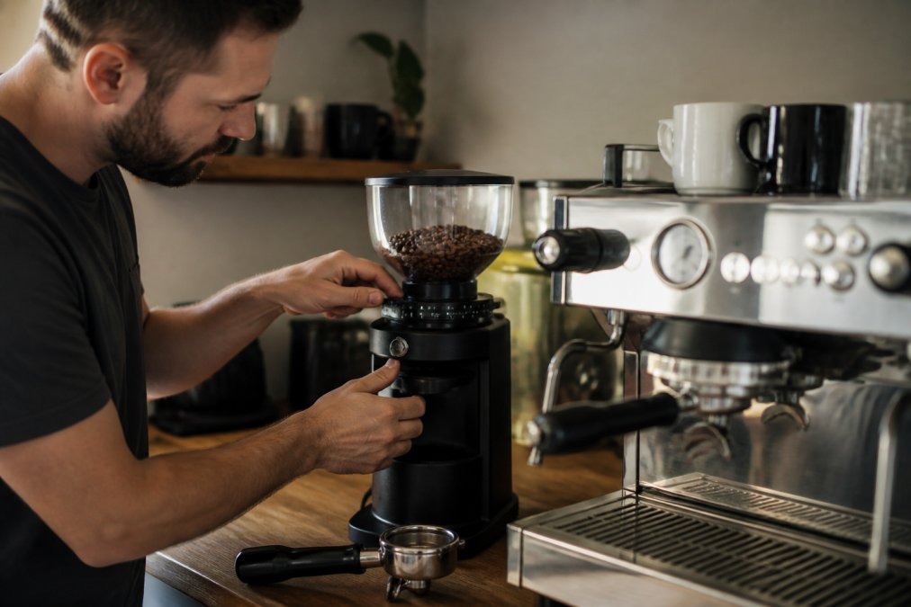 Home barista's hands adjusting the grind size dial on a burr grinder, with an espresso machine and portafilter visible in soft, warm kitchen lighting
