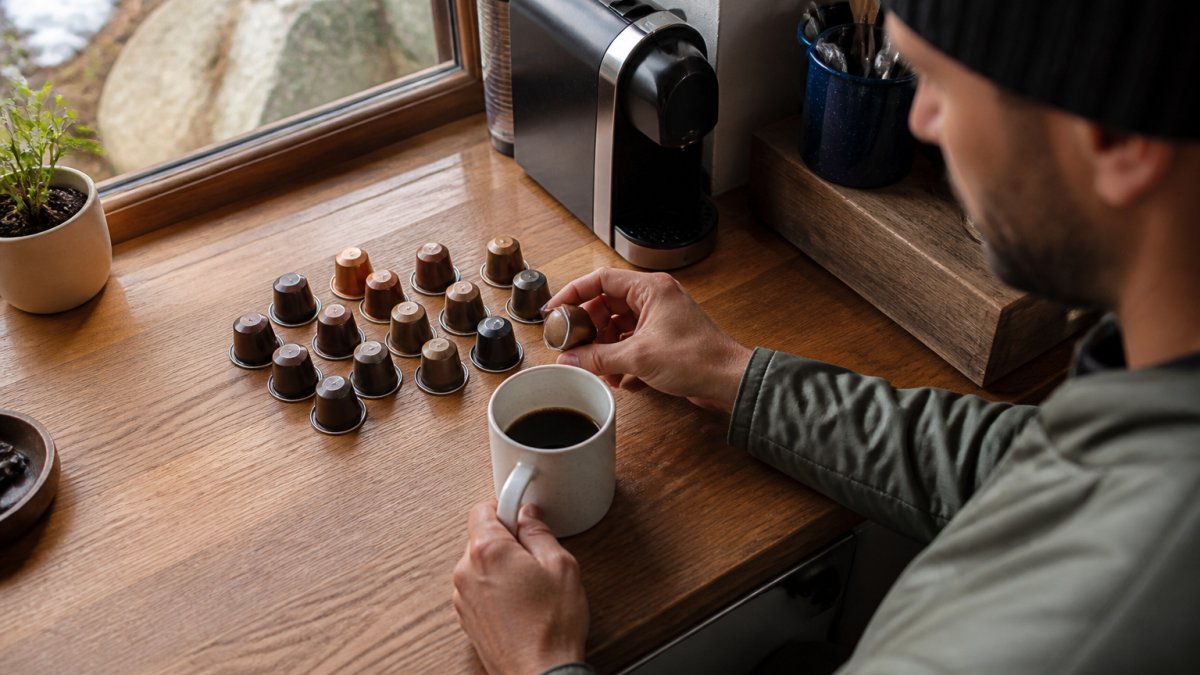 Overhead shot of several single-serve coffee pods in different roast shades arranged on a wooden break room counter next to a white ceramic mug, morning light through a window