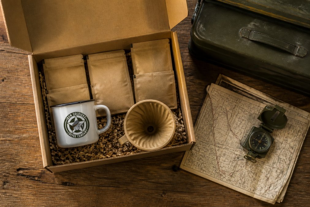 Overhead flat-lay of an open kraft subscription box on a weathered wooden table, revealing several 2oz sample coffee pouches, a vintage-style enamel mug, and a pour-over dripper, with a folded 1940s-style field map beside the box in warm natural light